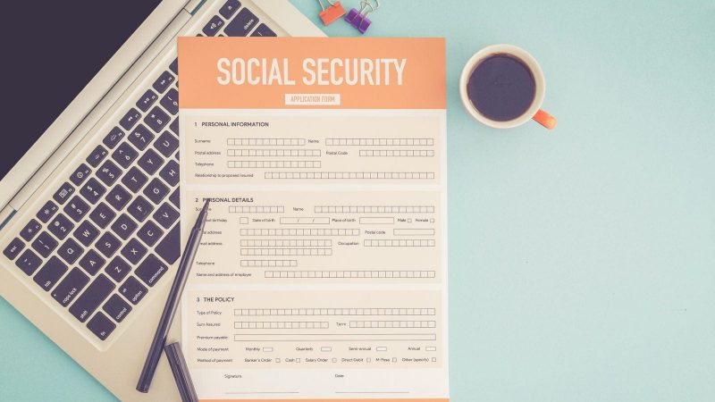 A Social Security application form on top of a laptop, on a desk with a cup of coffee and pens nearby.